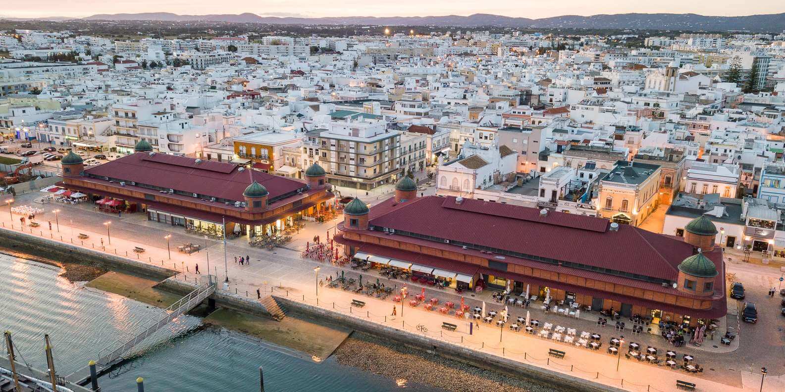 Vibrant waterfront walkway in Olhão featuring traditional red brick market halls, outdoor café seating, and clear views over the Ria Formosa lagoon